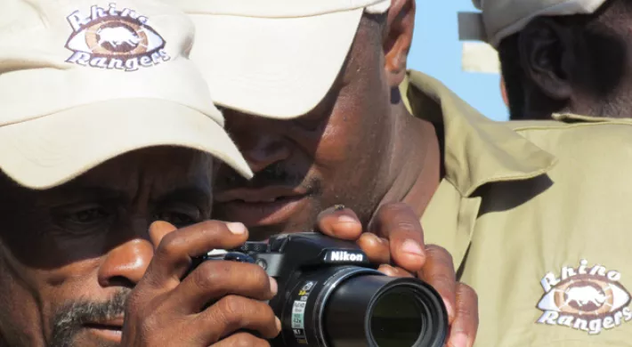 Two dark skinned men stand close together, staring at a digital camera screen. They are wearing tan baseball caps and tan buttons up shirts that say "Rhino Rangers" on the lapel. The back of another man's head is visible in the background.