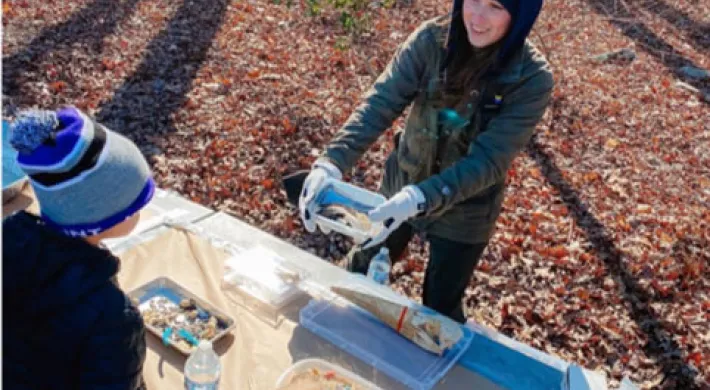 A woman in a navy hoodie and green jacket is standing at a table in a forest, smiling down at two children. She holds a plastic container and appears to be showing them its contents. The ground is covered in brown leaves and the long shadows of trees are visible on the ground.