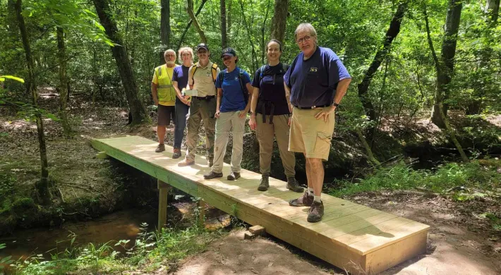 Trail building team consisting of six people of varying ages, races and genders, standing on a bridge that spans a small creek in the woods. They are standing in a line and posing for a photo.