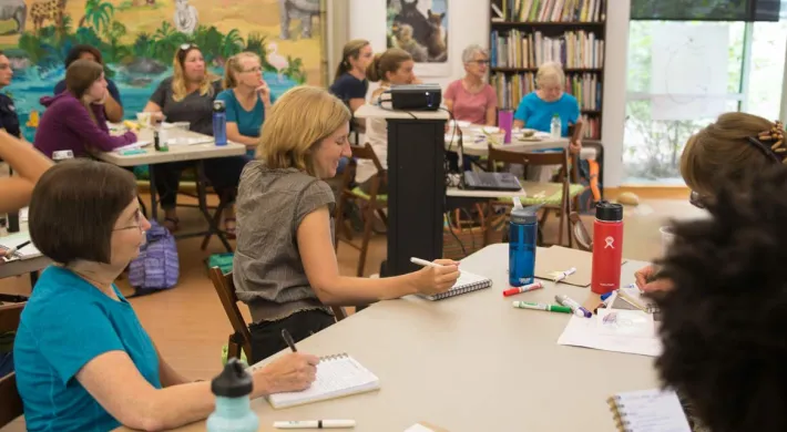 A group of about fifteen adults, mostly women, are seated at light-colored tables in a bright room, facing a woman in a blue shirt who is standing and gesturing towards a projected image on a white board. Colorful animal artworks, including a large polar bear and a macaw, adorn the walls behind the standing woman.