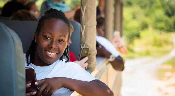 A group of kids sit in large bus seats on the back of an open air safari truck. The focus is on a young person with shoulder length dark braids, who is staring at the viewer, smiling while holding a cellphone in one hand and leaning an elbow out of the side of the truck.