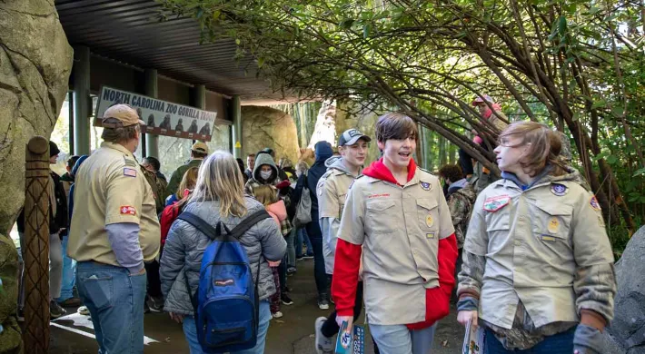 A large group of people with varying ages gathered in front of and walking by a large window and rock wall. They are all wearing Boy Scout uniforms consisting of jeans and tan collared shirts with patches on them.
