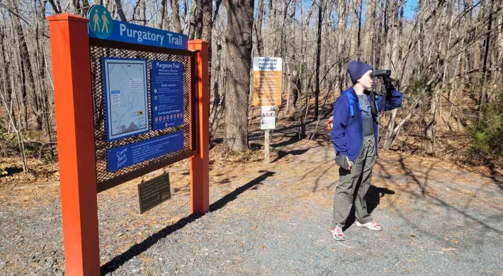 A person in cold weather gear, stands on a trail in the woods, looking off to the right with a pair of binoculars. Next to them is a large sign with the title "Purgatory Trail" with a map and some instructions underneath.