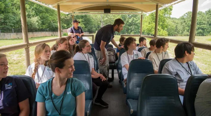 A group of students viewing animals from the open-air Zoofari bus as part of their Wild Science Camp experience. It is driving through a large, open field and trees are visible in the background.