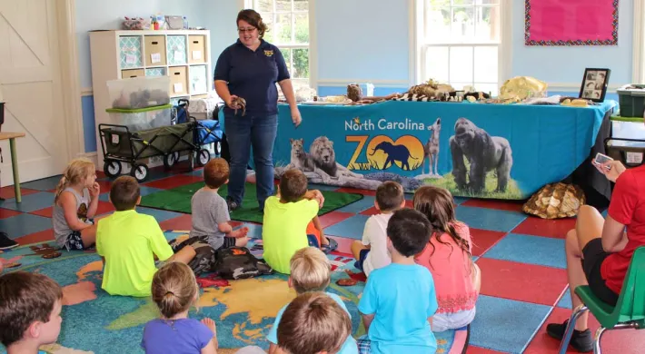 A woman in a navy NC Zoo uniform stands with a long table, covered with a teal NC Zoo tablecloth and various animal artifacts. In front of her is a group of children seated on colorful floor tiles, and a woman with a long blonde ponytail and red collared shirt sitting in a green chair to the right. The setting appears to be an elementary school classroom.