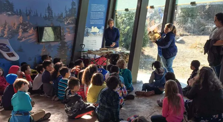 A group of approximately twenty children is sitting on the floor in a educational space with slanted glass windows. Two adult educators are leading the activity; one stands near a table with materials on the left, and another is kneeling to the right, showing the children an animal hide or fur. Large murals with landscape images and informational text are visible on the back walls.