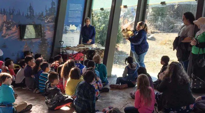 A group of approximately twenty children is sitting on the floor in a educational space with slanted glass windows. Two adult educators are leading the activity; one stands near a table with materials on the left, and another is kneeling to the right, showing the children an animal hide or fur. Large murals with landscape images and informational text are visible on the back walls.