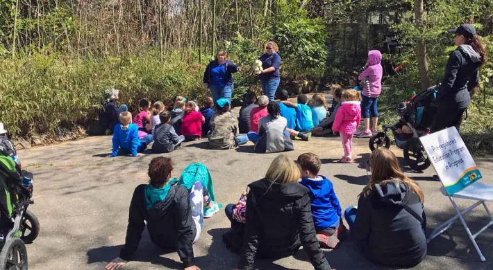 A group of children and adults sitting in a circle on a patch of dirt outdoors, possibly for a class or nature program. The people are dressed in various brightly colored jackets and shirts (including blues, reds, and pinks). The setting is a wooded area with green foliage in the background. A few people in the foreground have their backs to the camera, and some backpacks are visible.