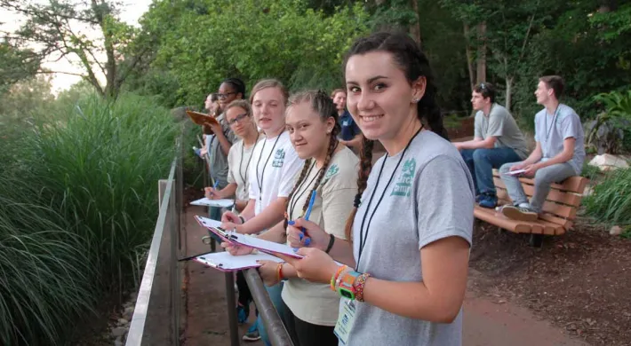 Seven students standing on a walkway outdoors, looking over a railing. The students are wearing matching gray or white t-shirts, some with lanyards, holding clipboards and pencils, appearing to be taking notes. The young woman in the foreground is smiling at the viewer. Tall, dense green grasses are on the left, and a few other students are seated on a wooden bench on the right, with a dirt path in front of them. The background is a mix of trees and foliage.