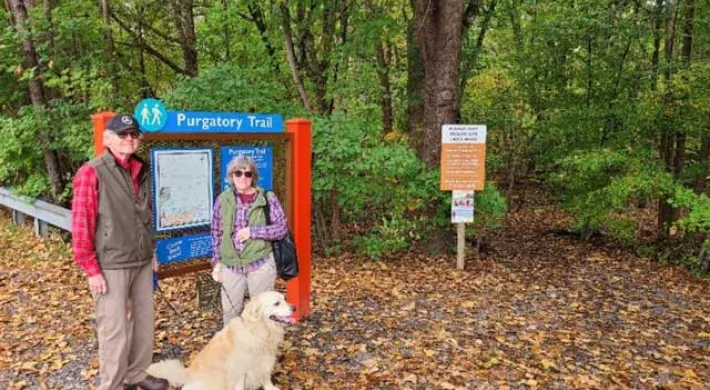 An older man and woman with their golden retriever pose beside a bright blue and orange trailhead sign for the Purgatory Trail on a leafy autumn path.