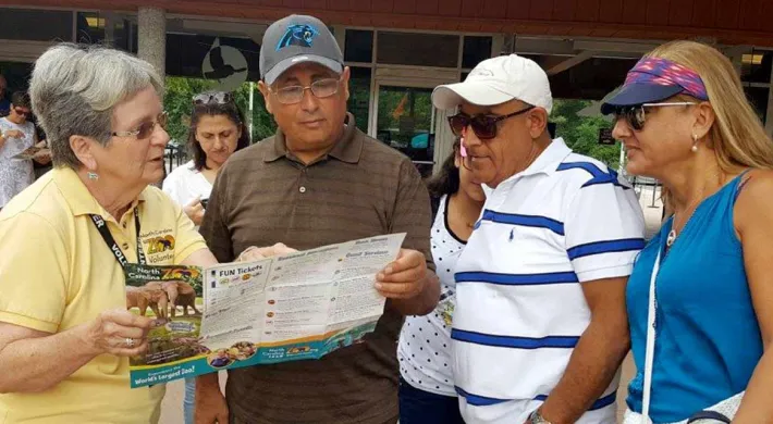 A zoo or park employee in a yellow shirt is helping three visitors by pointing to information on a folded map or brochure. They are standing outside near the entrance to a building.