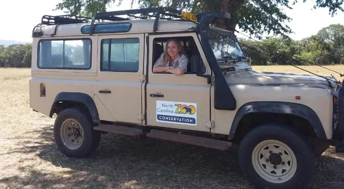 A light tan Land Rover Defender or similar safari-style off-road vehicle is parked in a sunny, dry field with trees in the background. A person with light hair is visible leaning out of the open driver's side door.