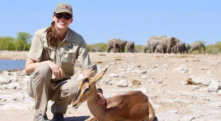 A woman in a tan rangers uniform squats smiling with a hand resting on the long delicate neck of an Addra Gazelle lying next to her. The two are on dry, rocky soil next to a small lake. A herd of Elephants can be seen in the far distance.