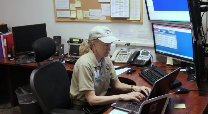 An adult man wearing a tan uniform shirt and a white baseball cap is seated at a desk in an office, working on a computer with multiple monitors.