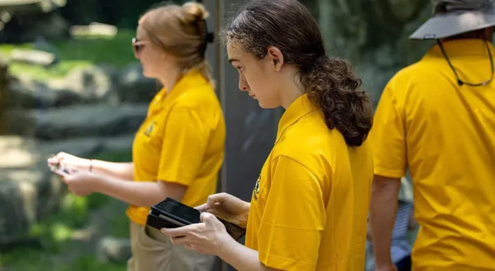 Three zoo or park employees wearing bright yellow uniform shirts are standing near an animal enclosure. The center person, with long, dark, curly hair, is looking down at a tablet or handheld device.