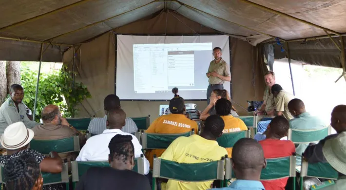 A large outdoor canvas tent, where a group of people are attending a presentation or training session. A man in a khaki shirt is standing under a projected screen on the back wall, speaking to the seated audience. About a dozen individuals, mostly men, are seated on metal-framed chairs, facing the front. 