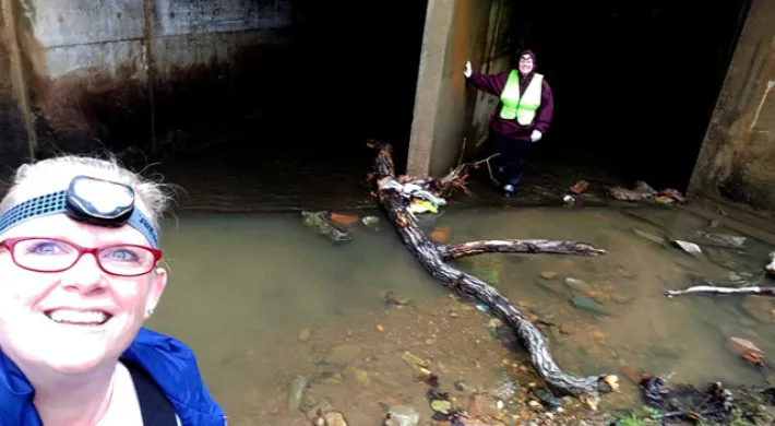 A low-angle selfie of two women standing in front of a dark concrete storm drain outlet. The woman closest to the camera is wearing a blue jacket, red glasses, and a headlamp, and is smiling. The second woman stands in the background, inside the dark entrance, wearing a dark jacket and a high-visibility safety vest. The ground is a shallow stream of muddy water containing rocks, trash, and a large piece of driftwood.