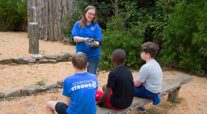 A nature educator, a woman with glasses and a blue uniform shirt, is holding a dark snake while presenting to three young boys seated on a wooden bench. The setting appears to be an outdoor educational area, possibly at a zoo or nature center, with a dirt ground and a wooden fence/structure in the background.