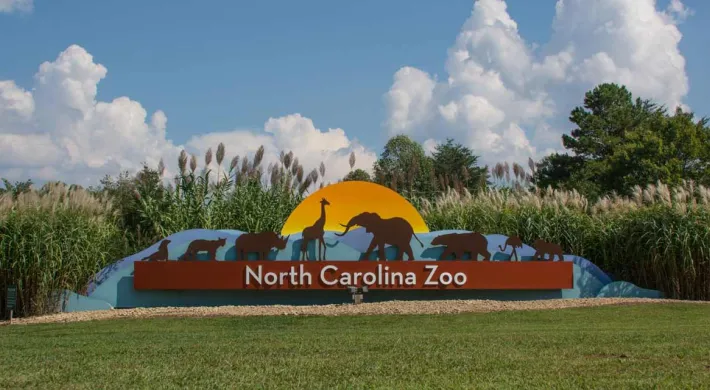 North Carolina Zoo Entrance Sign with animal silhouettes and colorful shapes and sun behind the animals.