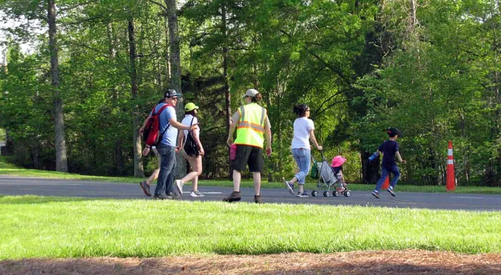 A diverse group of people is walking along a paved path, with a large, grassy area to their left and trees and bushes to their right. The group includes two adults pushing a stroller, a couple, and a man in a bright yellow safety vest who appears to be directing or monitoring the group.