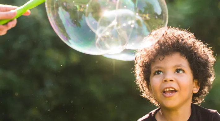A child with short, curly hair enjoying some time with giant bubbles outside in the sun.
