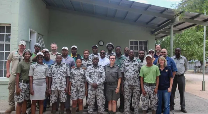 A large group of approximately 20 people, consisting of men and women, pose together for a group photo in front of a light green, single-story building. Most of the individuals are wearing matching camouflage or light-colored ranger/conservation uniforms, with a few wearing other casual outdoor attire. They appear to be a team of park rangers or conservation staff.
