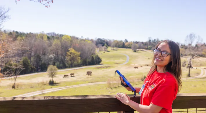 Research volunteer in a red shirt smiles at camera while overlooking the Watani grasslands habitat. Rhinos and antelope are on the hills in the background on a bright sunny day.