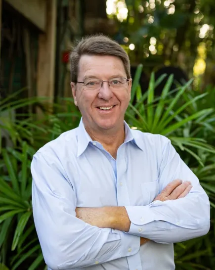 Doctor Mark Penning stands in front of a lush green backdrop of palm plants; he appears relaxed, with arms crossed, while wearing a light blue button down collared shirt and a beaming smile.