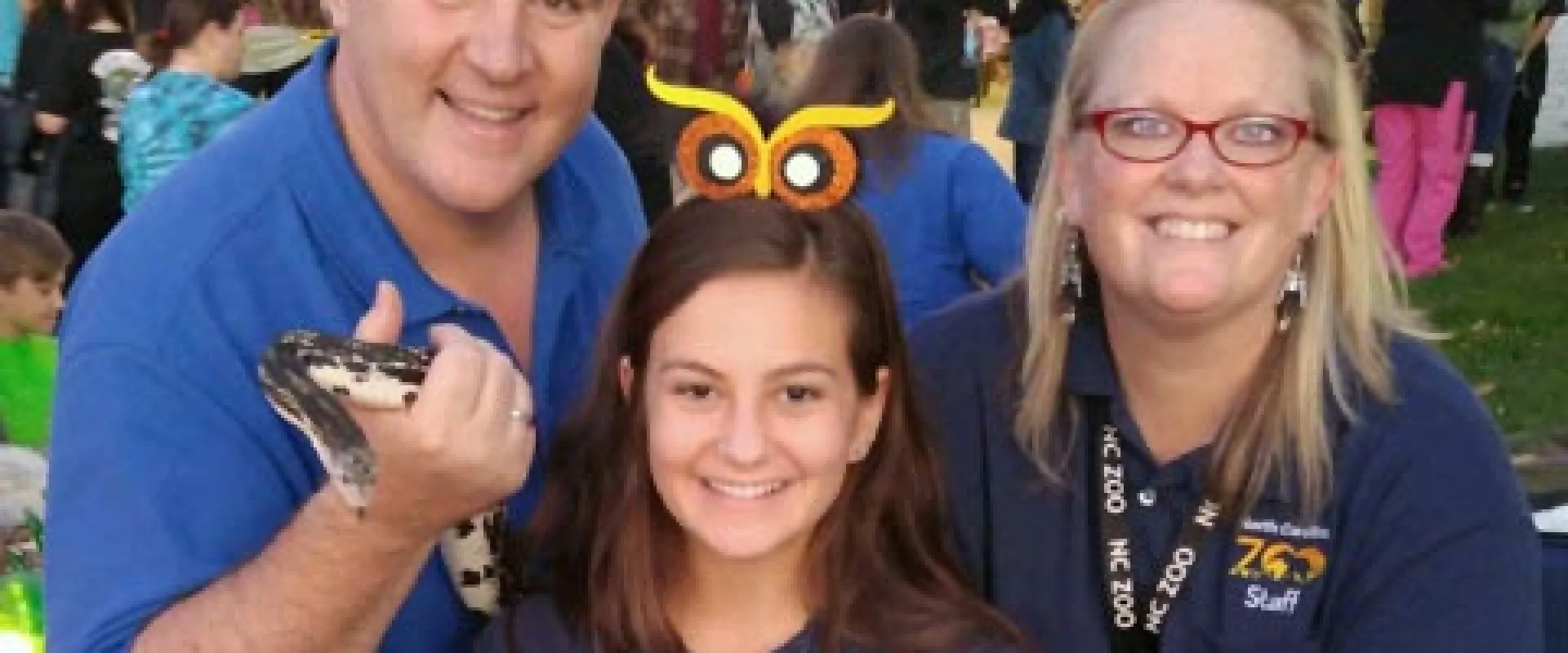 Three smiling community educators, one male and two female, holding various animal ambassadors, including a snake, tarantula, and two hissing cockroaches, at a Fall Festival event with crowds of eventgoers in the background.