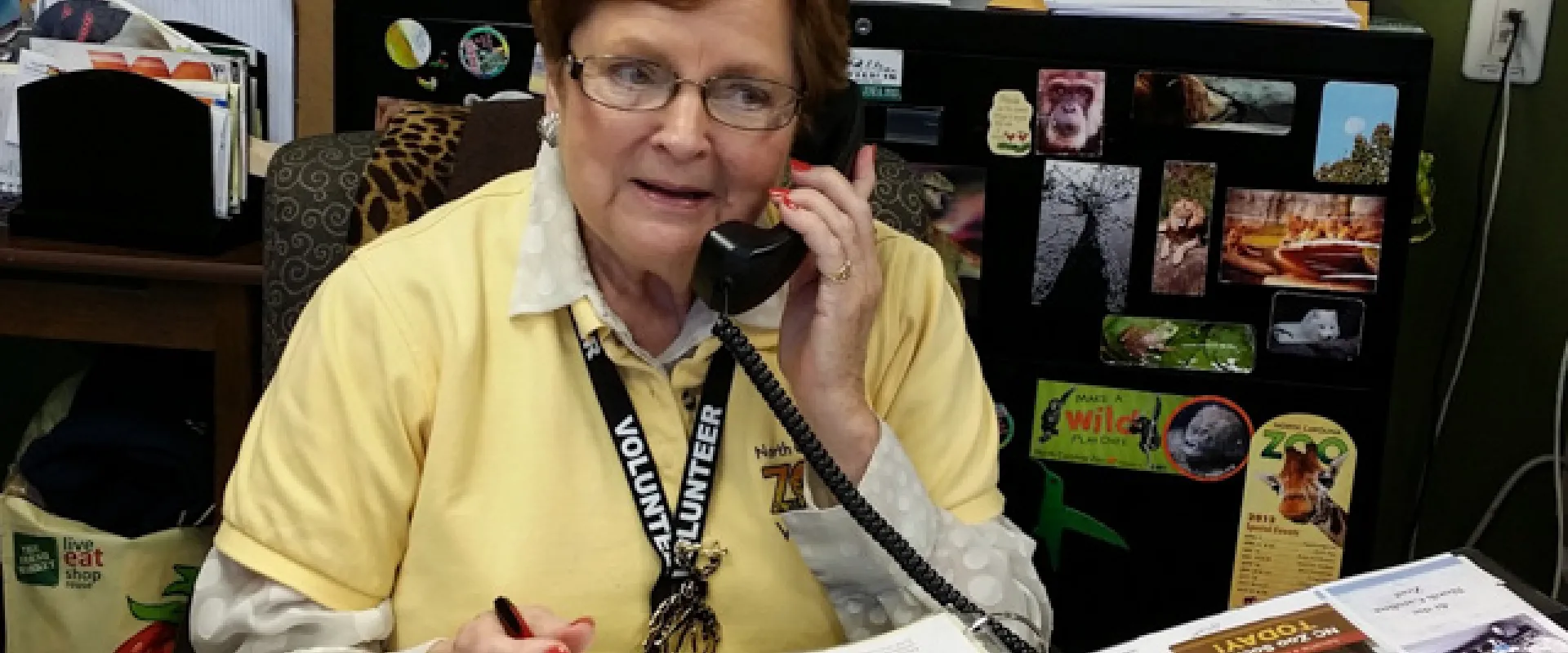 An older woman with glasses and short, red hair, wearing a yellow collared shirt, appears to be in an office talking on a corded telephone while sitting at a desk that is covered with papers. There is a black filing cabinet covered with magnets behind her.