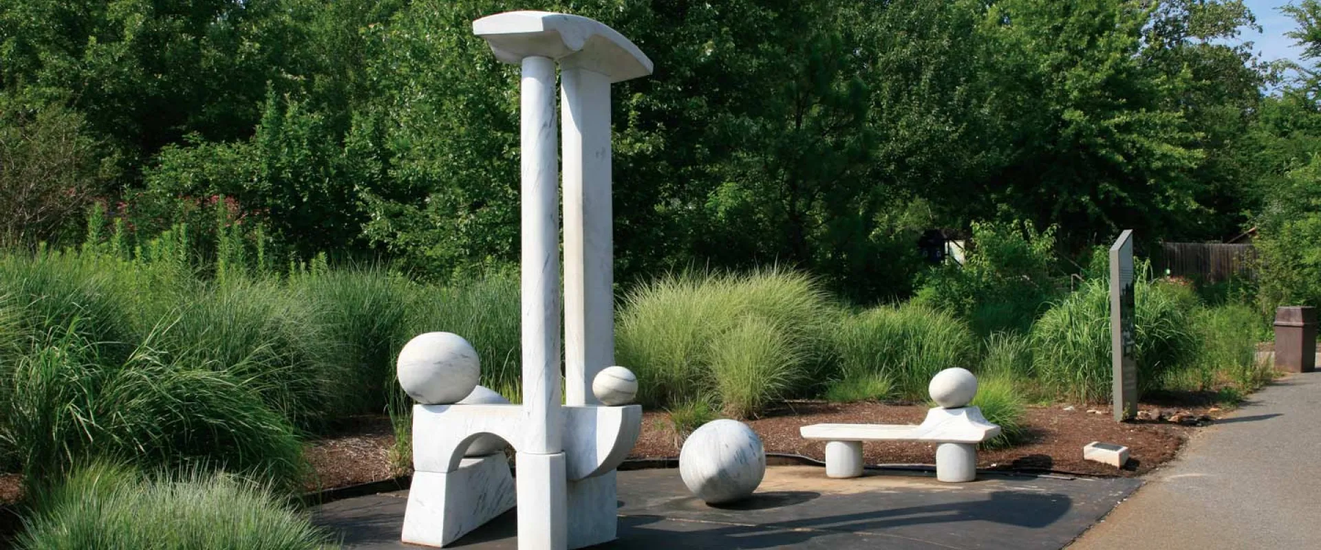 Zoological egg rest art installation which is comprised of minimalist white stone sculptures in a lush green outdoor setting, with a tall central piece, spherical elements, and a bench-like form beside a paved walkway.