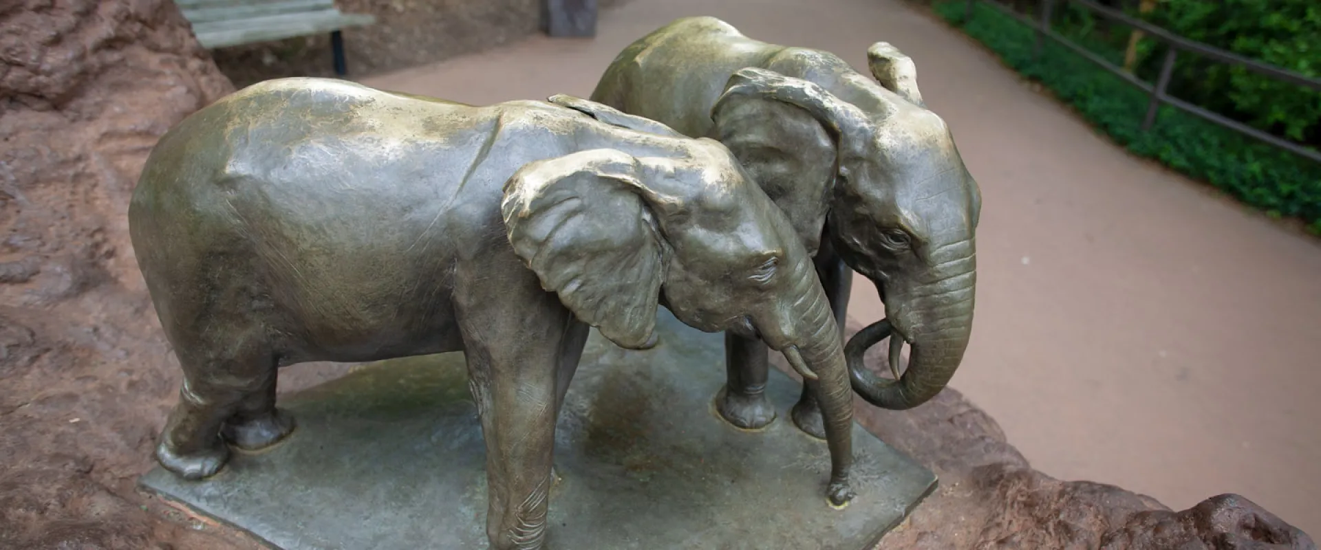 Small, bronze sculpture of two elephants standing together, one with their trunk curled while the other is straight, which are set atop a large brown rock.
