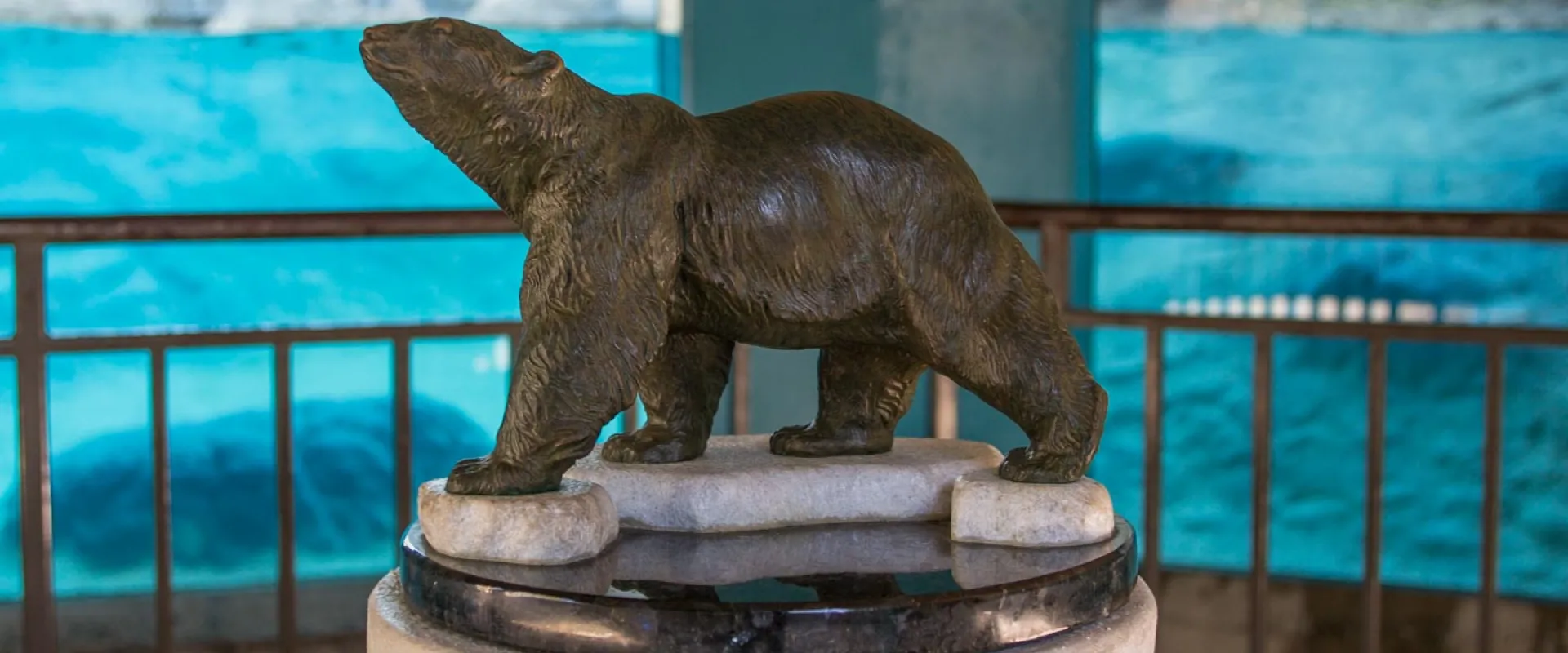A medium sized bronze statue of a Polar Bear walking on ice, rests on a large pilar in front of the polar bear habitat. There is a guardrail and blue water behind glass in the background.