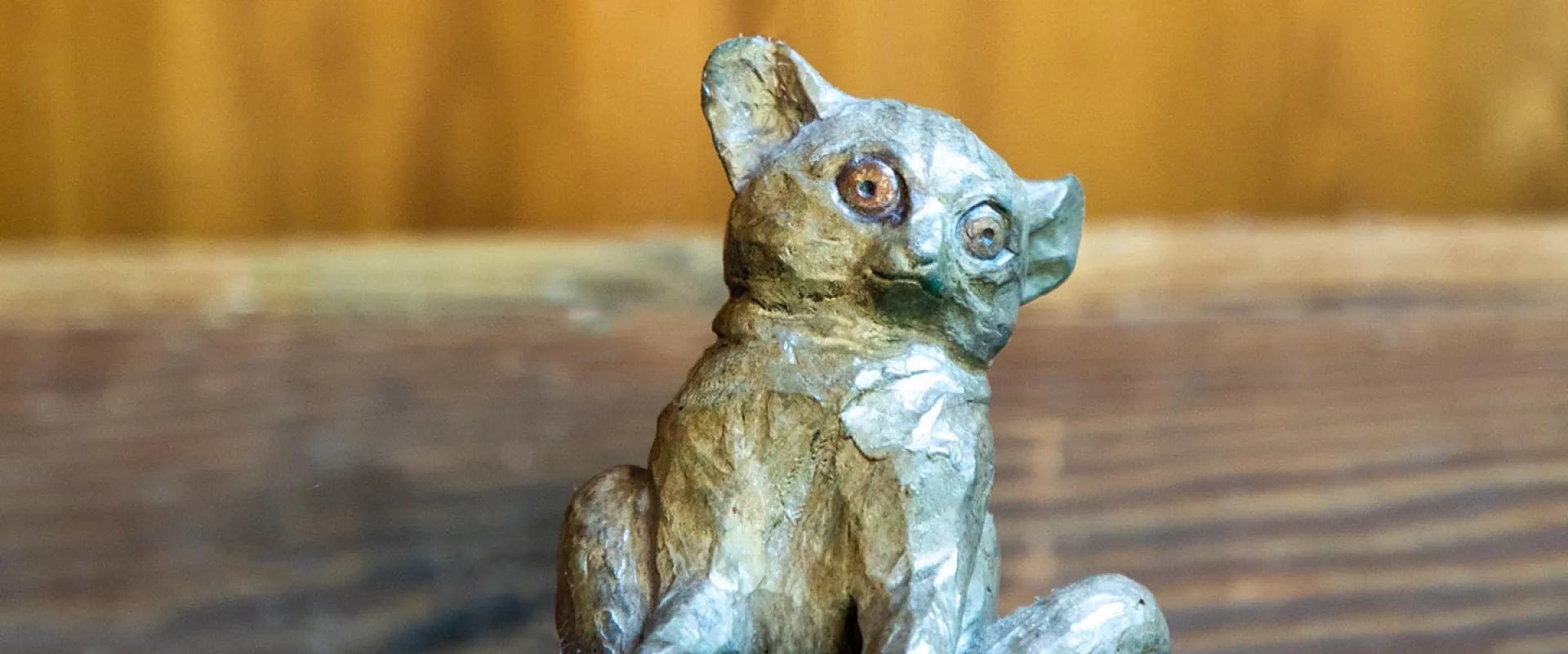 A small, bronze sculpture of a Bush Baby-a small, mousey looking primate with small, rounded ears and large, round eyes sitting in front of a wooden wall.