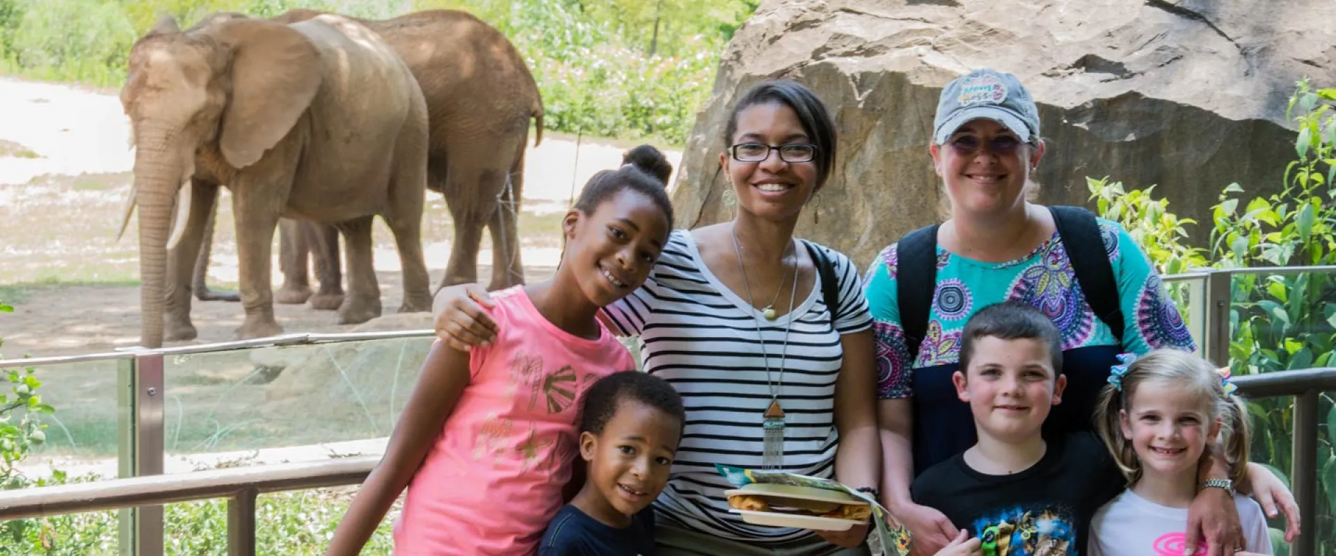 A group of six individuals, appearing to be a group of friends, pose for a photo in front of an elephant enclosure. In the background, two African elephants with their characteristic large ears and tusks stand near a rocky, grassy area. In the foreground, from left to right, a young girl with dark skin and her hair in a bun smiles broadly, wearing a pink shirt; a young boy with dark skin looks towards the camera; a woman with dark skin and glasses, smiles with her arm around the girl.