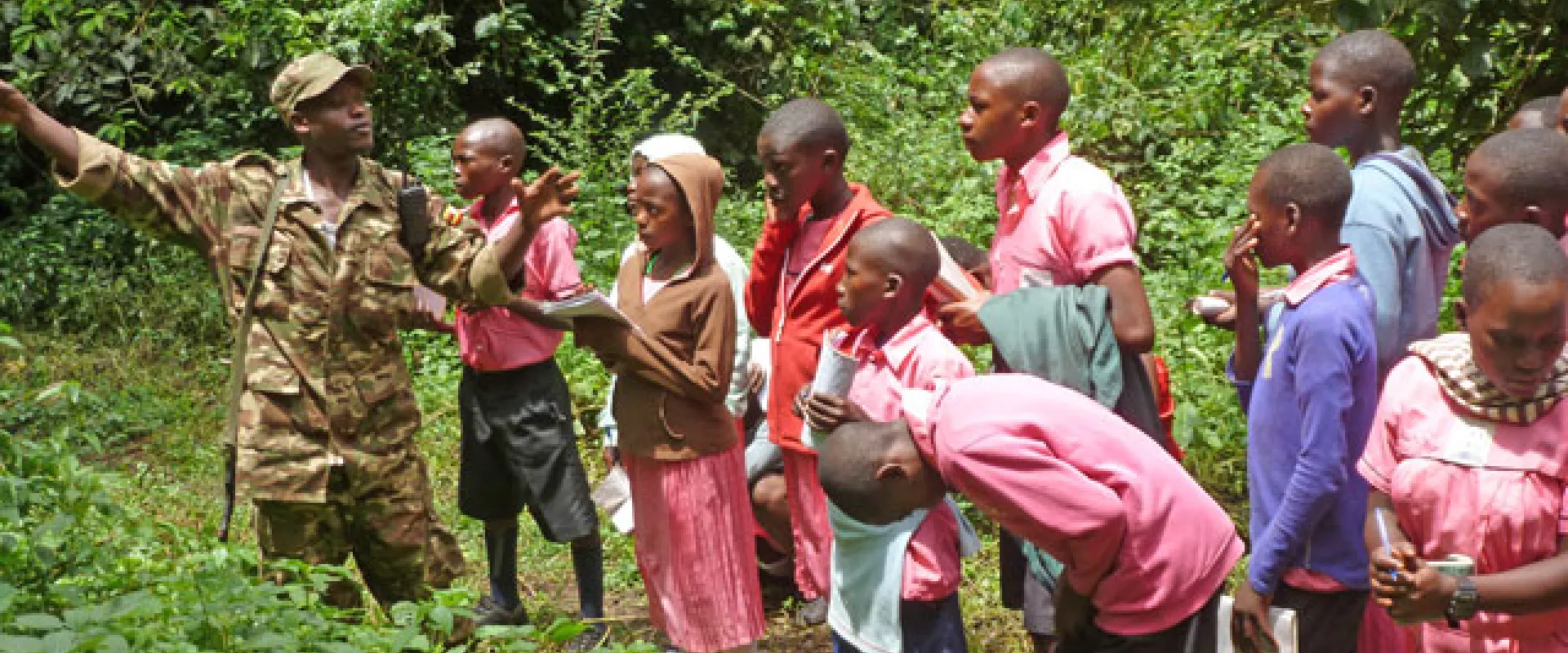 A group of children, most appearing to be Black, are gathered in a grassy area with dense foliage, listening to a person in a military-style uniform who is gesturing and pointing. The children are wearing various colored tops and dresses. The children are students from the UNITE program on a field trip.