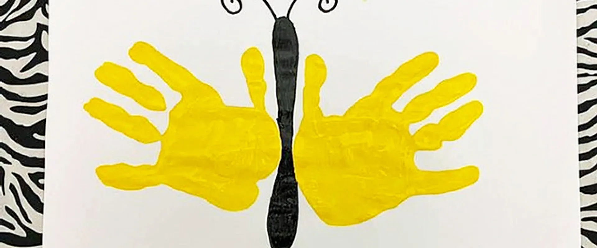 A Butterfly craft that is made by drawing a long, thin, brown body and antennae. The wings are made by making handprints with yellow paint. It is on a white paper background with a black and white Zebra striped border.