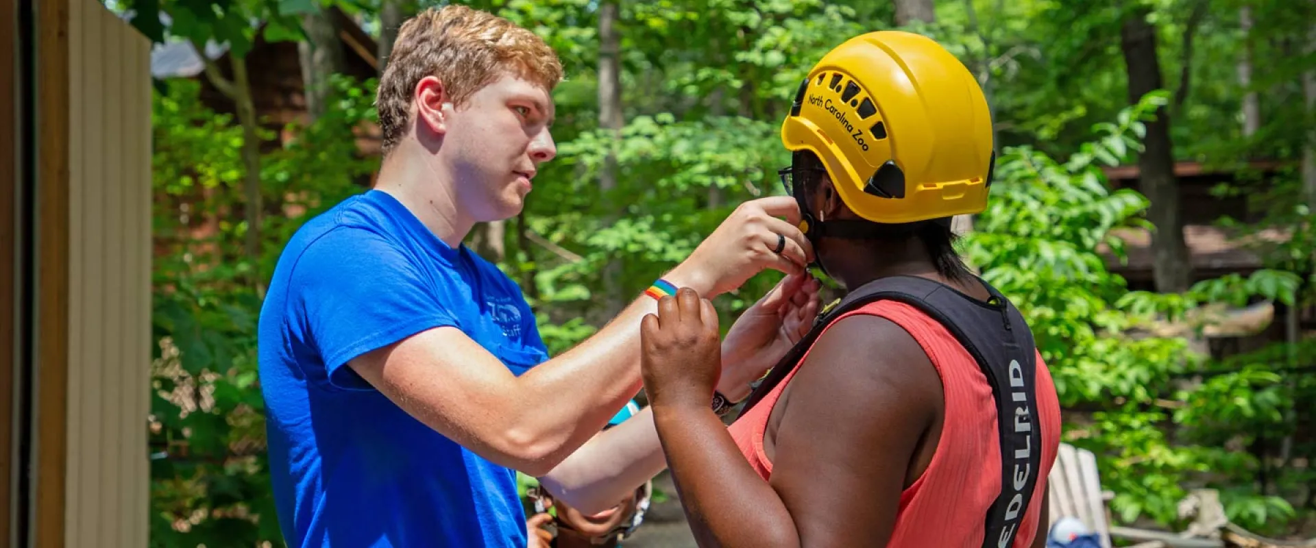 A young seasonal staff member is helping get a guest ready for Air Hike ropes course by fastening her yellow helmet beneath her chin. She is also wearing a black harness across her shoulders and back. They are standing outdoors in a wooded area,
