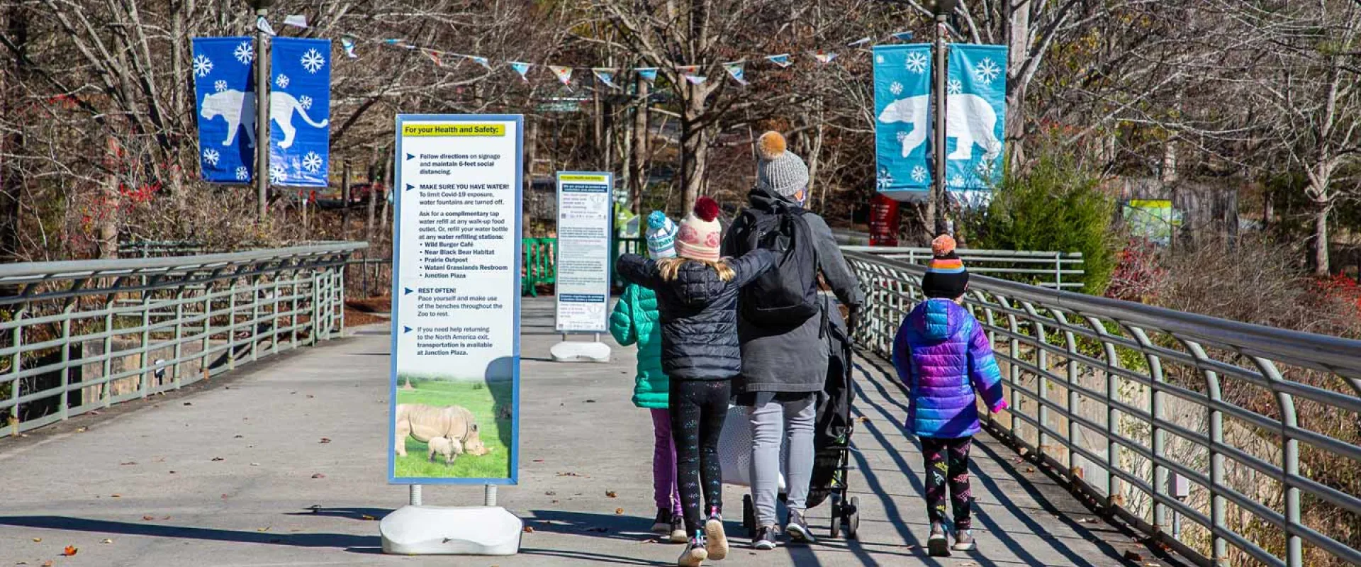 A group of Winter guests entering the Zoo. Three individuals including one adult and two kids walk over a large cement bridge that has tall handrails on either side and two tall informational signs placed at either end. In the background are trees, colorful pennant flags, and blue banners with pictures of Winter animals on them.