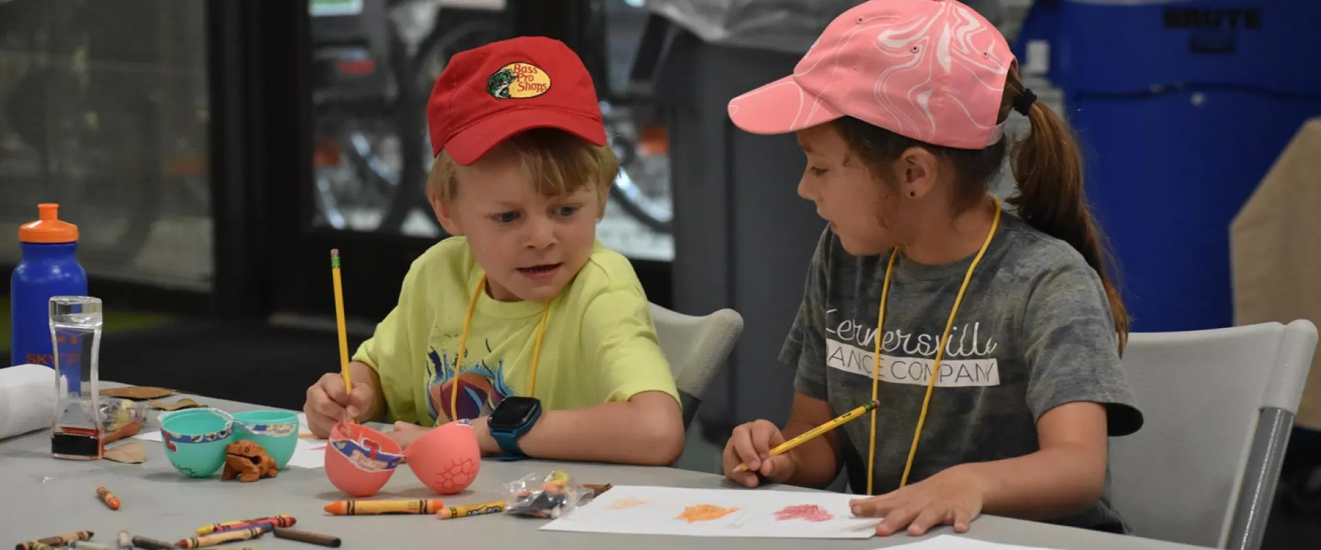 Two children sitting at a fold out table with paper, pencils, crayons, plastic eggs, and water bottles, doing a camp craft. The child on the left is a boy with  short blonde hair and a red baseball hat, and to the left is a girl with a dark pony tail and a pink baseball hat.