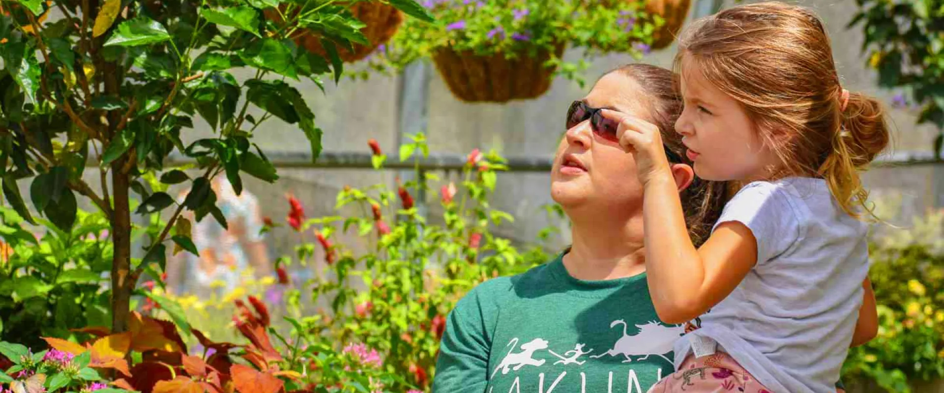 A woman wearing sunglasses and a green t-shirt holds a young girl with blonde hair in a ponytail. Both are looking upwards and seem to be observing something. They are surrounded by lush green and colorful plants, with hanging baskets visible in the background, suggesting they are in a garden or greenhouse.