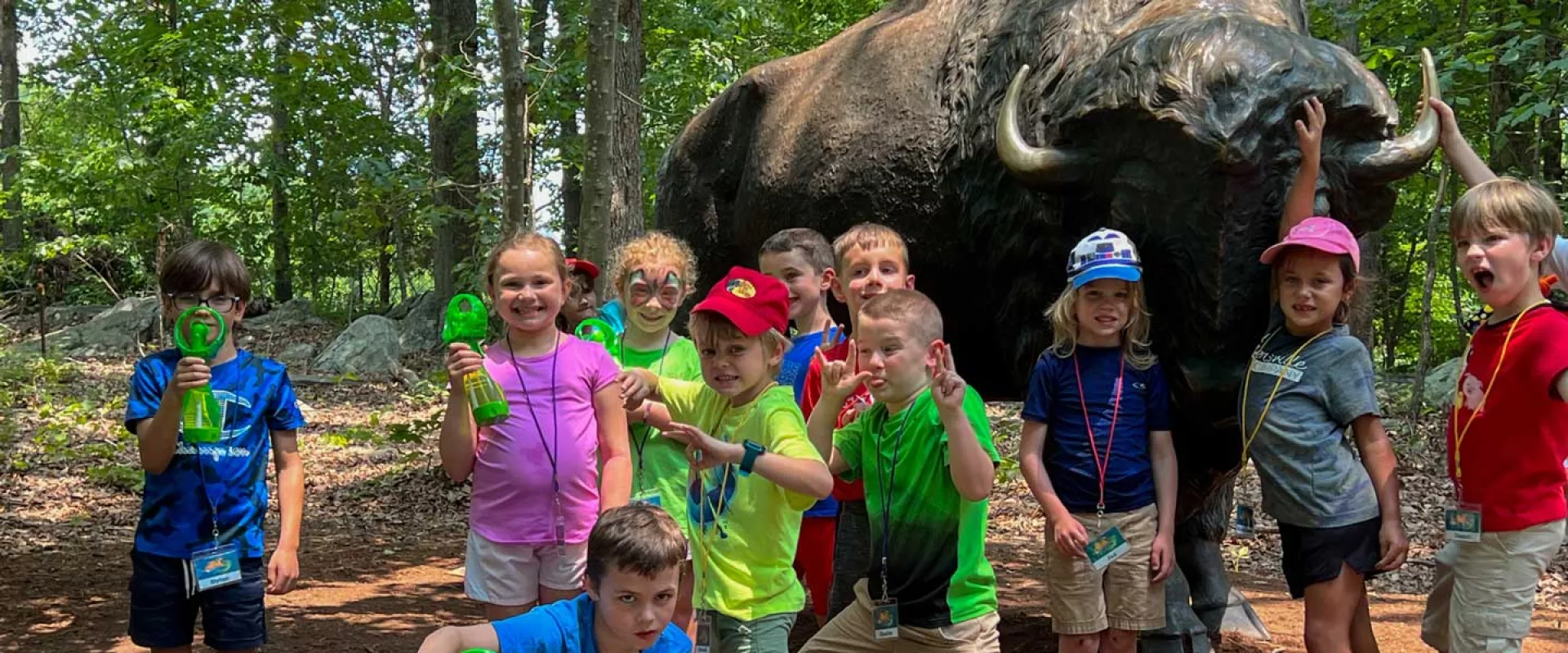 A group of young campers holding green personal fans stand together, posing in front of a large brass statue of a Bison in a wooded area.