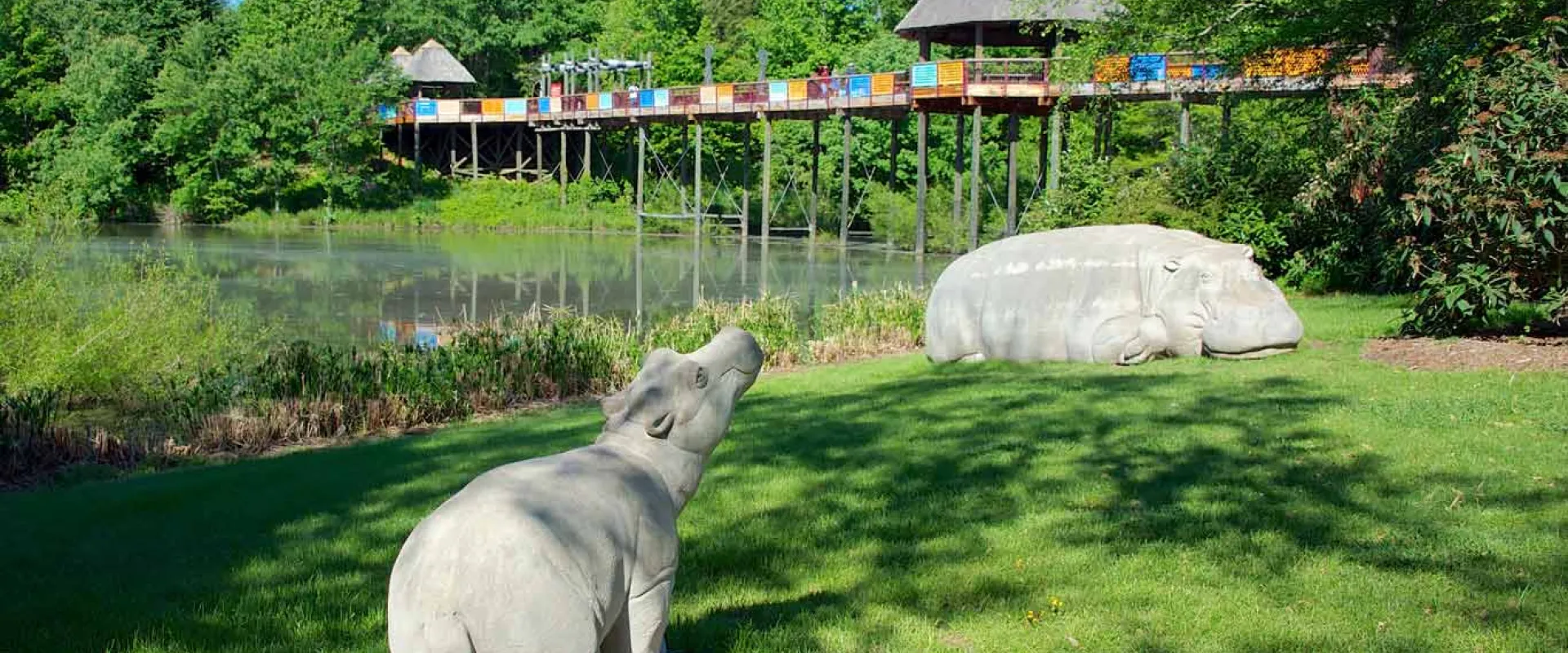 Two limestone hippos statues standing on the grassy shores of a large lake. A long wooden bridge with straw roofed gazebos at either end and colorfully painted railings, crosses the lake in the background. 