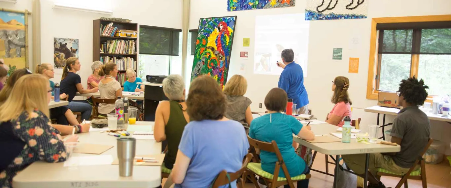 A group of about fifteen adults, mostly women, are seated at light-colored tables in a bright room, facing a woman in a blue shirt who is standing and gesturing towards a projected image on a white board. Colorful animal artworks, including a large polar bear and a macaw, adorn the walls behind the standing woman.