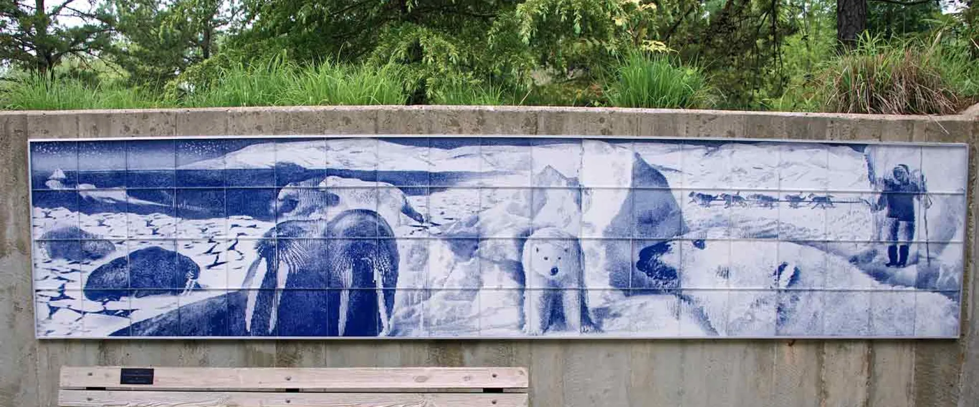 A wide, light wooden bench with a black metal frame sits in front of a long, rectangular mural on a concrete wall. The mural, rendered in shades of blue and white, depicts Arctic wildlife, including walruses and polar bears, and a human figure in winter gear. Green trees and foliage are visible above the wall in the background.