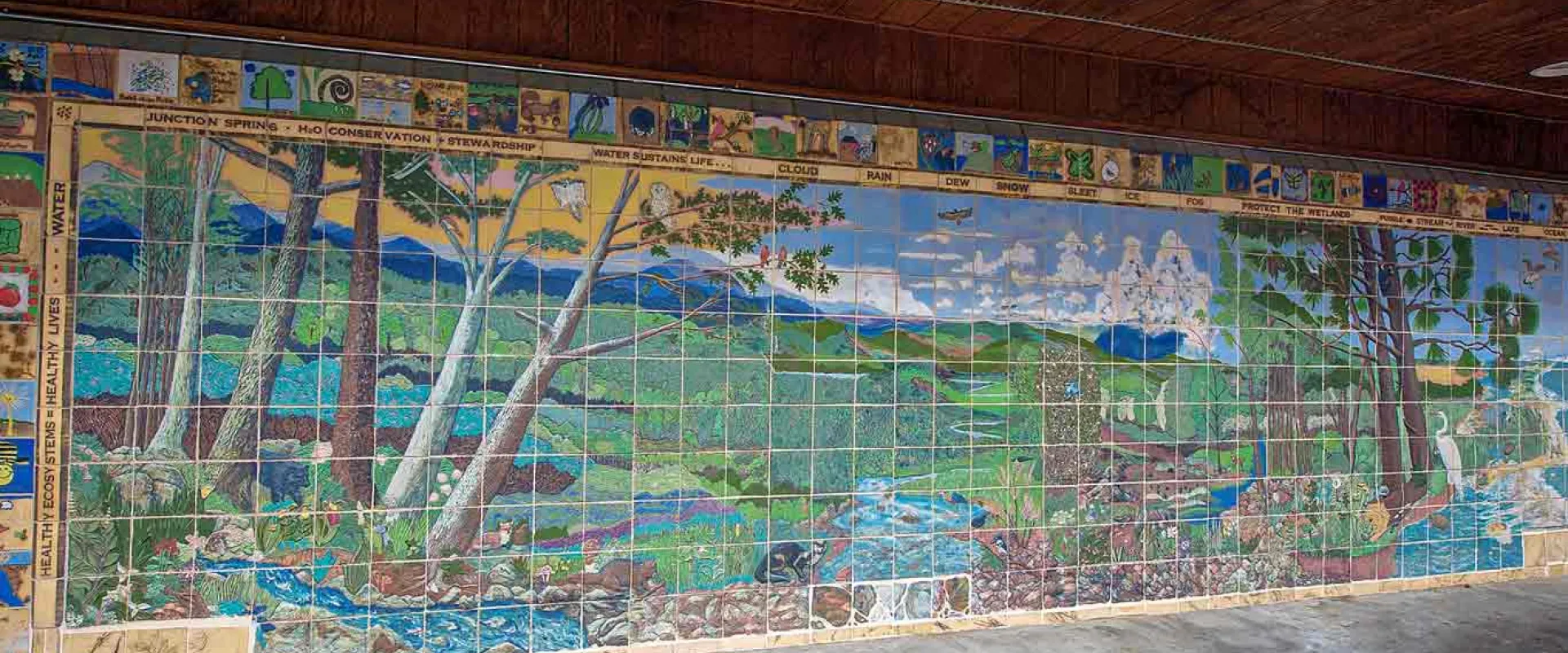 A vibrant, large-scale mural made of square tiles depicts a lush, expansive landscape. On the left, tall trees with light-colored trunks and green foliage frame a scene that transitions into a wide-open field in the center. A light blue river flows from the left foreground to the center. The mural extends across a long wall under a brown overhang.