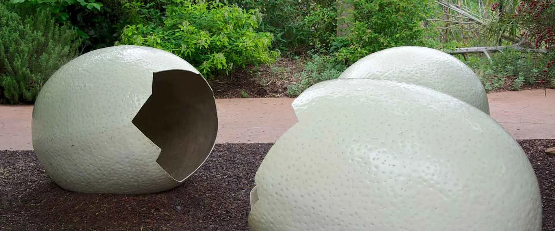 Artistic installation featuring three oversized, pale eggshell sculptures rest on dark earth, One eggshell on the left is visibly cracked open with jagged edges, revealing its hollow interior. The other two are mostly intact, showing a textured surface.