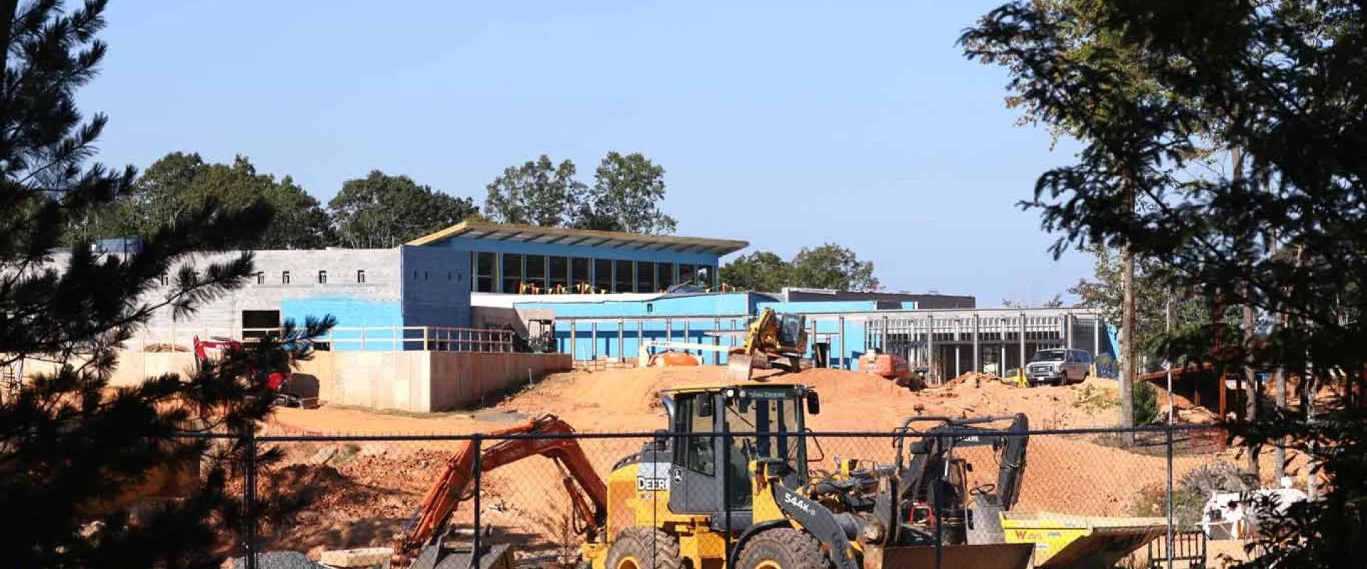A sunny, distant view of a construction site with multiple pieces of heavy machinery, including an excavator, on reddish-brown dirt. In the background, a light blue and grey building under construction is visible, surrounded by green trees under a clear blue sky, framed by dark foliage in the foreground.