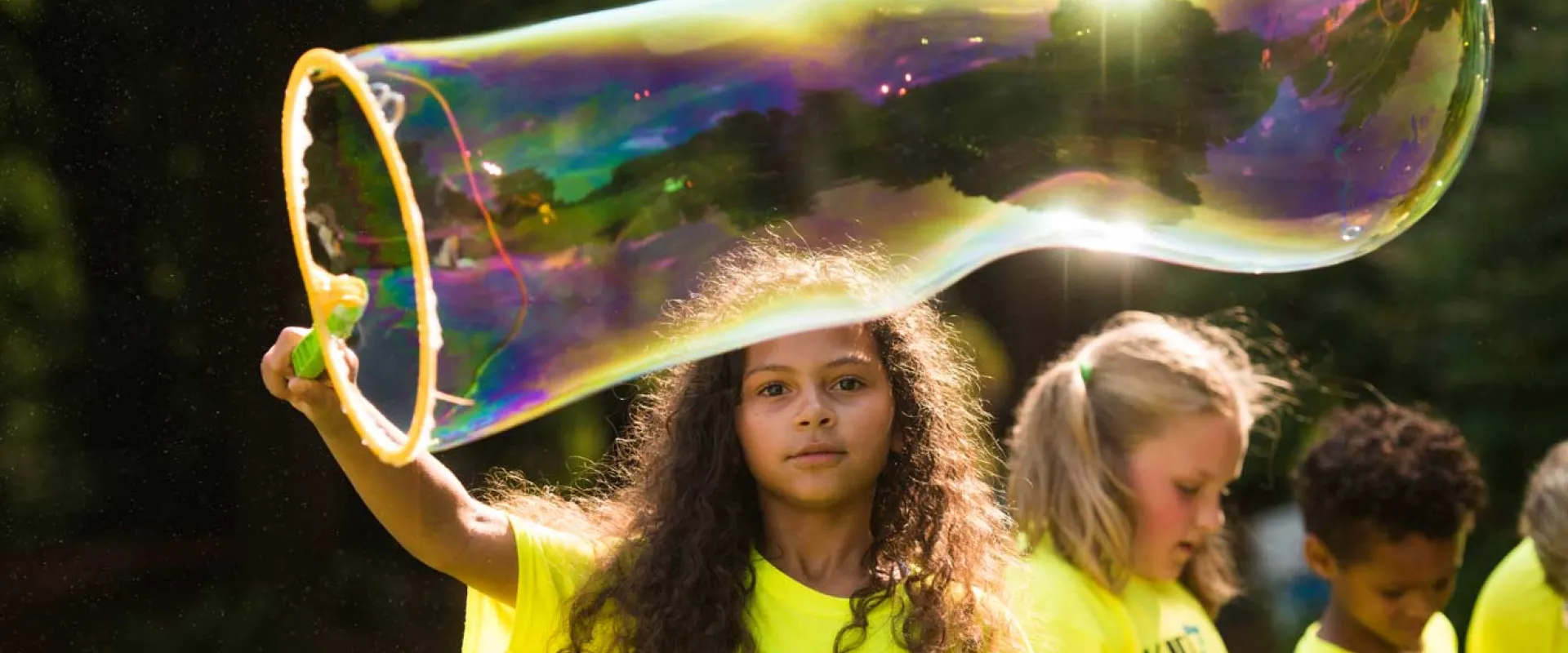 A young girl with long curly hair uses a wand to blow a giant bubble. There are trees and a group of other kids out of focus in the background.