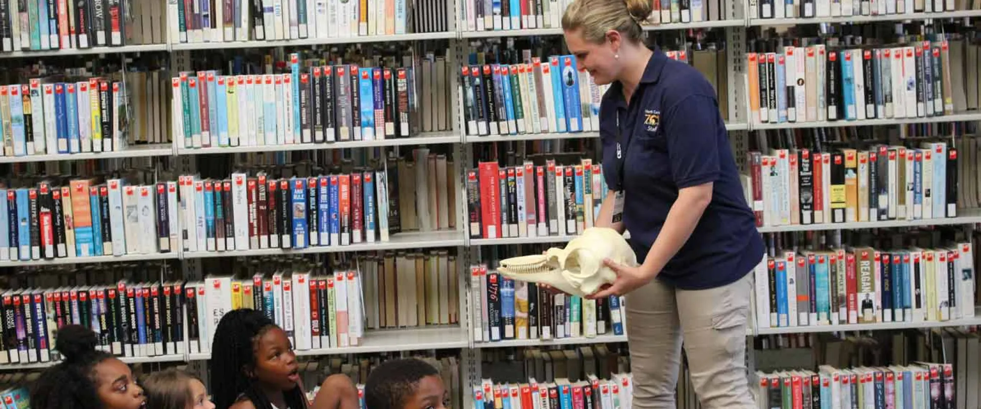 A woman with a blonde bun and navy NC Zoo uniform shirt stands in front of a group of children holding a large skull. A wall of bookshelves filled with books fills the wall beside her, giving the impression of an educational presentation at a school library.
